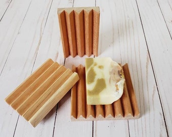 May include: Three wooden soap dishes with a natural finish. One dish holds a bar of soap with green and white tones. The dishes have a slatted design to allow for drainage. The soap dish is a bathroom accessory.