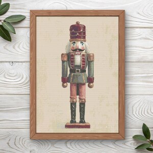 May include: A framed wall art print featuring a centered illustration of a nutcracker soldier. The figure wears a red hat, a green jacket with gold trim, and black boots. The background is a textured beige, and the frame is a medium-toned wood.