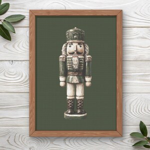May include: A framed cross-stitch pattern featuring a nutcracker soldier in a green uniform with gold accents, a tall hat, and white beard, centered on a dark olive green fabric background within a light brown wooden frame.