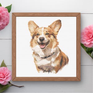 Op de afbeelding: Een ingelijst kruissteek borduurwerk met een portret van een Pembroke Welsh Corgi met beige en witte vacht, puntige oren en een geopende bek met een roze tong, tegen een witte achtergrond in een lichtbruine houten lijst.