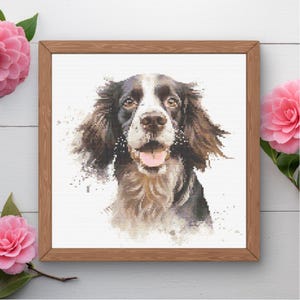May include: A framed cross-stitch portrait of a dog with brown and white fur, a pink tongue, and a happy expression. The dog's portrait is set against a white background, and the frame is made of wood.