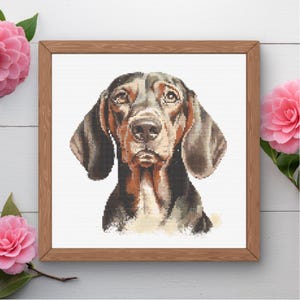 May include: A framed cross-stitch portrait of a Bloodhound dog. The dog has a brown and black coat, long floppy ears, and a soulful expression. The artwork is set against a white background and framed in a light brown wooden frame. Pink flowers are in the corners.