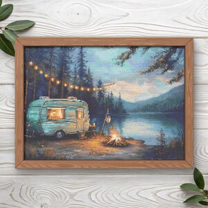 May include: A framed cross-stitch art piece depicting a teal vintage camper parked by a lake at dusk. A string of warm glowing lights hangs above the camper, and a small campfire burns nearby. The scene includes tall pine trees and mountains.