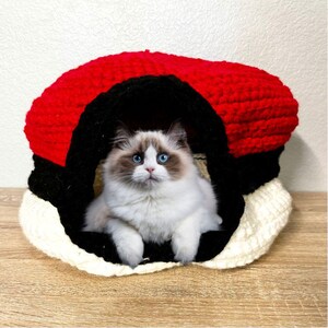 May include: A white cat with blue eyes sits inside a red, black, and white crocheted cat bed shaped like a Pokeball.