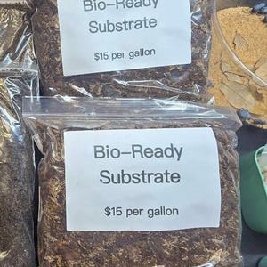 May include: Three clear plastic bags filled with dark brown organic substrate material. Each bag features a white rectangular label with the black printed text Bio-Ready Substrate and $15 per gallon. The contents appear to be a mix of soil and wood.