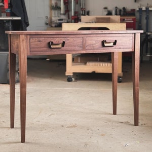 May include: A dark brown wooden console table with two drawers and brass pulls. The table has a simple, modern design and is made of solid wood. The table is standing on a concrete floor in a workshop setting.