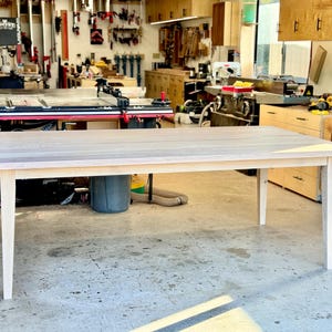 May include: A white wooden table with a rectangular top and four legs. The table is in a workshop setting with tools and other furniture in the background.