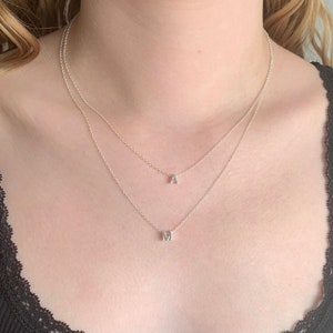 May include: A silver necklace with two chains and two small pendants, one with the letter 'A' and the other with the letter 'M'.