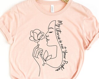 Peut inclure: Un t-shirt rose pâle avec un dessin au trait noir d'une femme qui sent une rose. Le texte "My Uterus is not your Business" est écrit sur le t-shirt.