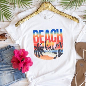 Może przedstawiać: Biała koszulka z nadrukiem graficznym przedstawiającym zach&oacute;d słońca z palmami i tekstem "Beach Bum". Tekst jest w tęczowym gradiencie kolor&oacute;w.