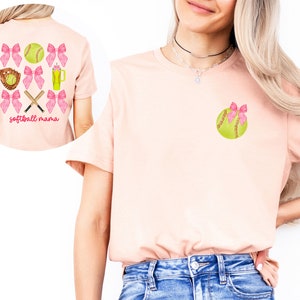 May include: A pink t-shirt with a softball design on the front and back. The front design features a single softball with a pink bow. The back design features a softball, bats, a glove, and a drink with pink bows around each item. The text "softball mama" is printed below the design.