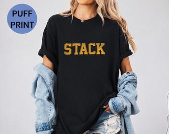 Pode incluir: Camiseta preta com a palavra "STACK" impressa em letras douradas.