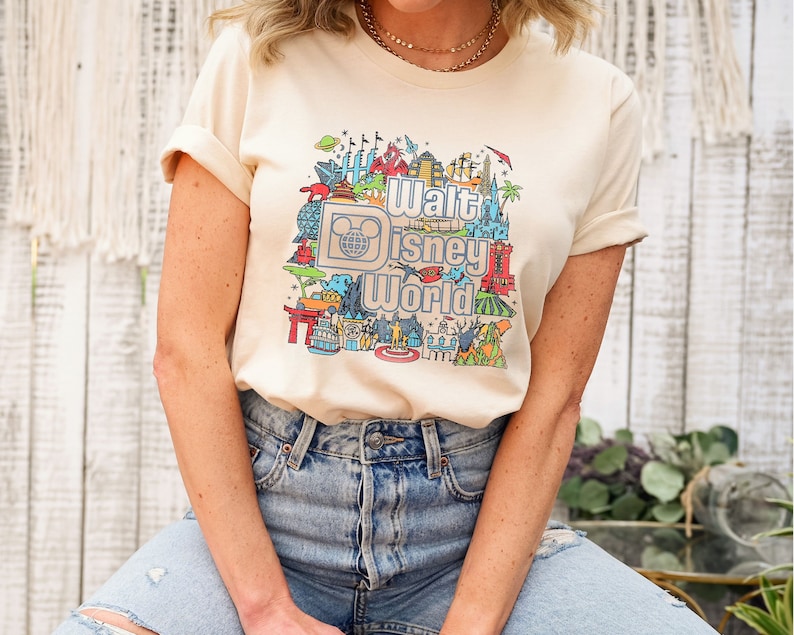May include: A beige t-shirt featuring a colourful graphic print of Walt Disney World landmarks and the text "Walt Disney World".