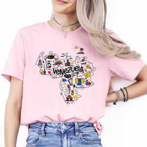 Może przedstawiać: R&oacute;żowa koszulka z grafiką przedstawiającą zarys Wenezueli z kolorowymi ilustracjami zabytk&oacute;w i symboli. Tekst "Venezuela" jest napisany czarnymi literami.