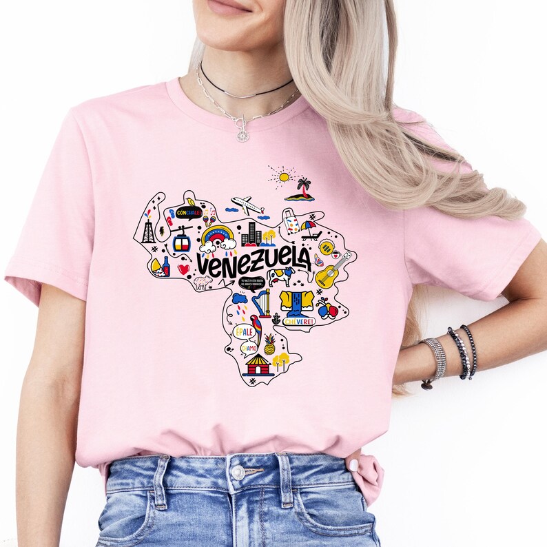 Może przedstawiać: R&oacute;żowa koszulka z grafiką przedstawiającą zarys Wenezueli z kolorowymi ilustracjami zabytk&oacute;w i symboli. Tekst "Venezuela" jest napisany czarnymi literami.
