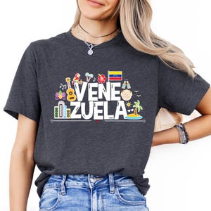 May include: A dark gray t-shirt with a white graphic design that says "Venezuela" and features various colorful icons representing the country, including a parrot, a guitar, a palm tree, and a flag.