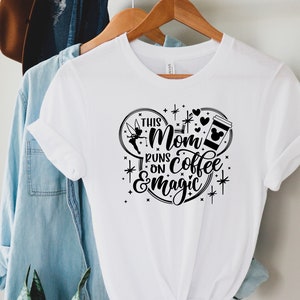 May include: White t-shirt with a black graphic design of a Mickey Mouse ear silhouette. The text inside the silhouette reads "This Mom Runs on Coffee & Magic" with a Tinkerbell fairy graphic and a coffee cup with Mickey Mouse ears.