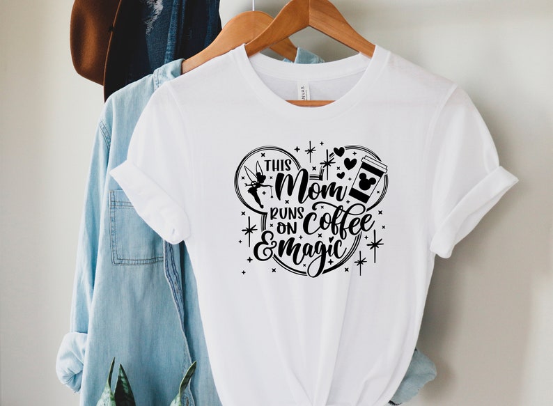 May include: White t-shirt with a black graphic design of a Mickey Mouse ear silhouette. The text inside the silhouette reads "This Mom Runs on Coffee & Magic" with a Tinkerbell fairy graphic and a coffee cup with Mickey Mouse ears.