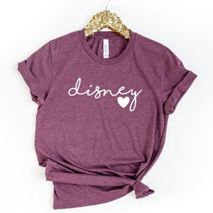 May include: A maroon t-shirt with the word "disney" in white script and a white heart graphic. The shirt is hanging on a gold sequined hanger. The sleeves are cuffed.