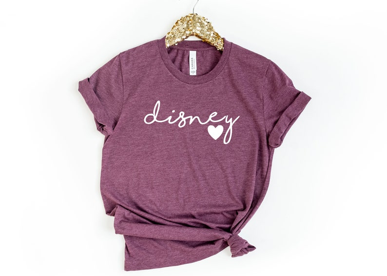 May include: A maroon t-shirt with the word "disney" in white script and a white heart graphic. The shirt is hanging on a gold sequined hanger. The sleeves are cuffed.