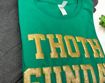 Pode incluir: Camiseta verde com letras douradas de glitter que dizem "Thoth Sunday".