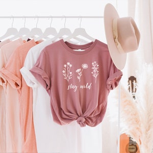 May include: A dusty rose t-shirt with a white floral design and the text "stay wild". The shirt is hanging on a white rack with other t-shirts in various colors.