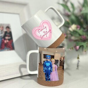 May include: Two white ceramic mugs with cork bottoms. The top mug has a pink heart with the text "Family is Everything". The bottom mug has a photo of two children in costumes.