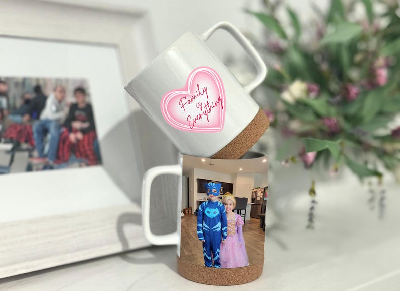 May include: Two white ceramic mugs with cork bottoms. The top mug has a pink heart with the text "Family is Everything". The bottom mug has a photo of two children in costumes.