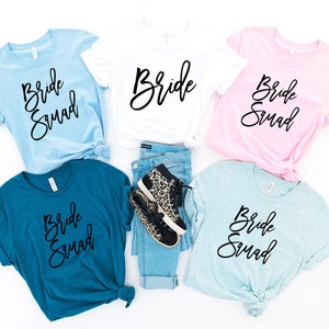 May include: Five t-shirts with the text "Bride Squad" in black script on different colored shirts. The shirts are laid out on a white surface with a pair of leopard print shoes and blue jeans.
