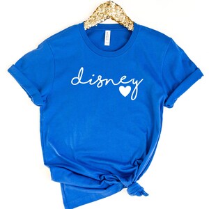 May include: A royal blue t-shirt with the word "disney" in white script and a white heart. The shirt is folded at the bottom and has rolled-up sleeves. The shirt is hanging on a gold sequined hanger.