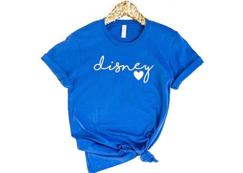 May include: A royal blue t-shirt with the word "disney" in white script and a white heart. The shirt is folded at the bottom and has rolled-up sleeves. The shirt is hanging on a gold sequined hanger.