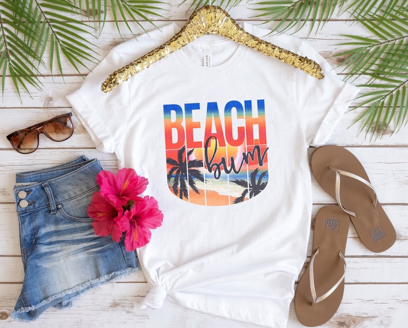 Może przedstawiać: Biała koszulka z nadrukiem graficznym przedstawiającym zach&oacute;d słońca z palmami i tekstem "Beach Bum". Tekst jest w tęczowym gradiencie kolor&oacute;w.