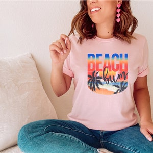 Może przedstawiać: R&oacute;żowy T-shirt z graficznym wzorem przedstawiającym zach&oacute;d słońca z palmami i tekstem "Beach Bum".