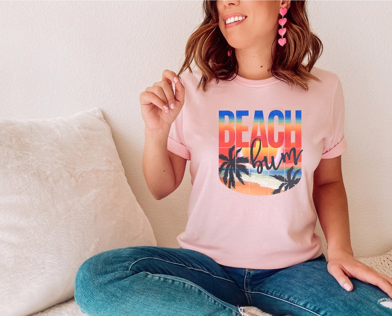 Może przedstawiać: R&oacute;żowy T-shirt z graficznym wzorem przedstawiającym zach&oacute;d słońca z palmami i tekstem "Beach Bum".