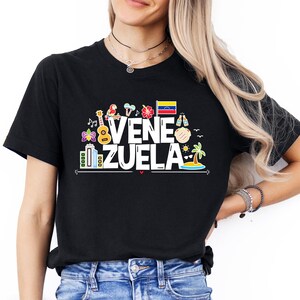 May include: Black t-shirt with a white graphic design featuring the word "VENEZUELA" surrounded by colorful icons, including a guitar, a parrot, a flag, and a palm tree.