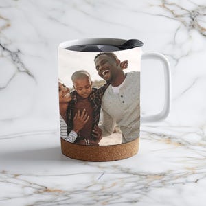 May include: White travel mug with a cork band and a photo of a family of three on the side. The photo shows a man, a woman, and a young boy smiling.