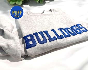 Könnte beinhalten: Grauer Pullover mit einem blauen Glitzer-Puffdruck, auf dem "BULLDOGS" steht.