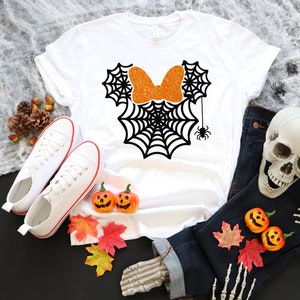 Op de afbeelding: Wit T-shirt met een zwart spinnenwebontwerp in de vorm van Mickey Mouse oren. De strik is van oranje glitter.