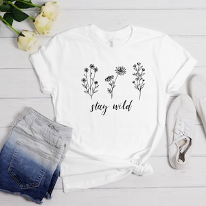 May include: White t-shirt with a black floral design and the text "stay wild". The shirt is laid out on a white wooden surface with a pair of white sneakers and denim shorts.