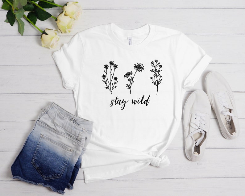 May include: White t-shirt with a black floral design and the text "stay wild". The shirt is laid out on a white wooden surface with a pair of white sneakers and denim shorts.