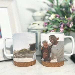 May include: Two white ceramic mugs with cork bottoms. One mug features a photo of a father and son smiling at the beach. The other mug features a photo of a person with long brown hair looking at the beach.