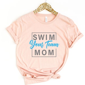 K&ouml;nnte beinhalten: Ein rosa T-Shirt mit dem Text "SWIM YOUR TEAM MOM" in einem grauen Kasten. Der Text "YOUR TEAM" ist in blau.