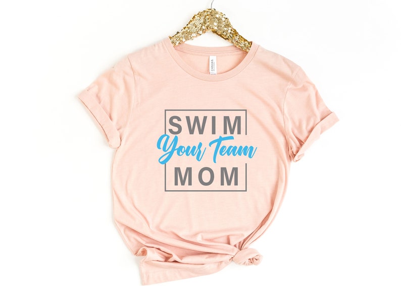 K&ouml;nnte beinhalten: Ein rosa T-Shirt mit dem Text "SWIM YOUR TEAM MOM" in einem grauen Kasten. Der Text "YOUR TEAM" ist in blau.