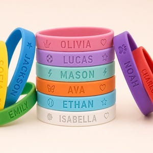 May include: A collection of colorful silicone wristbands, each engraved with a name and a small icon. The bands are in various colors, including pink, purple, blue, green, yellow, orange, and white. The names are in a sans-serif font.
