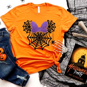 Op de afbeelding: Oranje T-shirt met een zwart spinnenwebontwerp en een paarse glitter strik. Het ontwerp lijkt op de oren van Mickey Mouse. Het shirt ligt op een wit oppervlak met een zwart-wit spinnenweb, een blauwe spijkerbroek en een Halloween-partij uitnodiging.