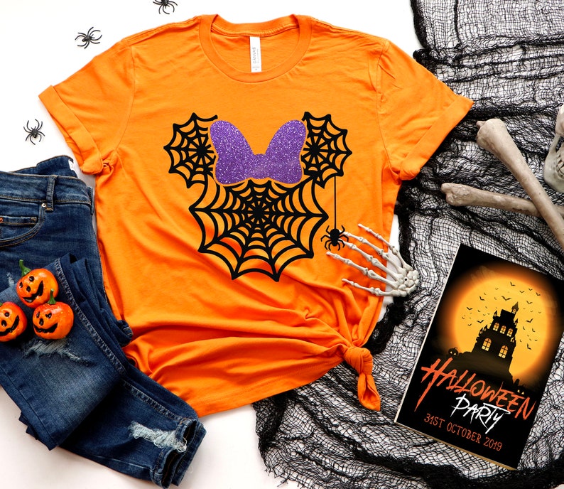 Op de afbeelding: Oranje T-shirt met een zwart spinnenwebontwerp en een paarse glitter strik. Het ontwerp lijkt op de oren van Mickey Mouse. Het shirt ligt op een wit oppervlak met een zwart-wit spinnenweb, een blauwe spijkerbroek en een Halloween-partij uitnodiging.