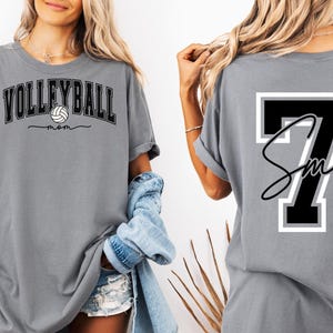 May include: Gray t-shirt with the text "VOLLEYBALL MOM" and a volleyball graphic on the front. The back of the shirt has a black number 7 with a white outline and the text "Smith" in cursive.
