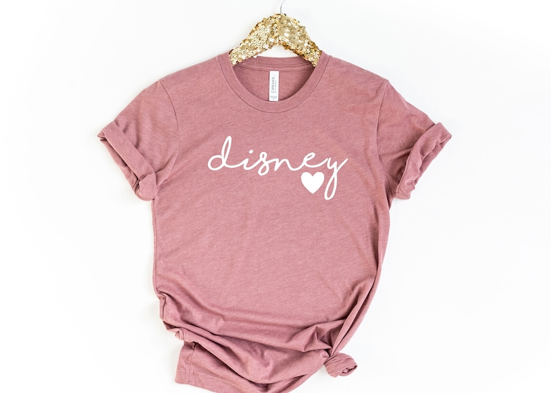 May include: A mauve-colored t-shirt with the word "disney" in white script and a white heart graphic. The shirt is hanging on a gold sequined hanger.