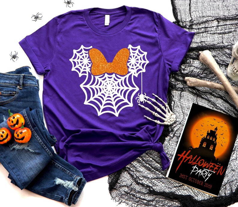 Op de afbeelding: Paars T-shirt met een wit spinnenwebontwerp in de vorm van Minnie Mouse oren. De strik is van oranje glitter. Het shirt ligt op een witte achtergrond met een blauwe spijkerbroek, een skelet hand en een Halloween-partyuitnodiging.
