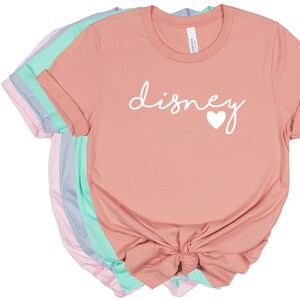 May include: A stack of t-shirts in various pastel colors, including pink, blue, and mint green. The front shirt is peach-colored and features the word "disney" in white script with a white heart. The shirts are folded with rolled sleeves.